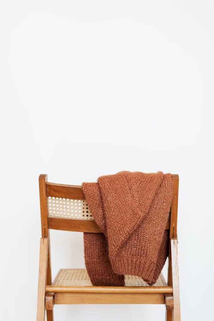 Brown coarse wool sweater hung on back of woooden chair against white wall